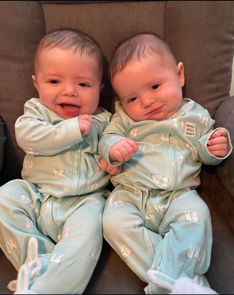 Identical twin babies sitting side by side, the twin on the right tilting his head.