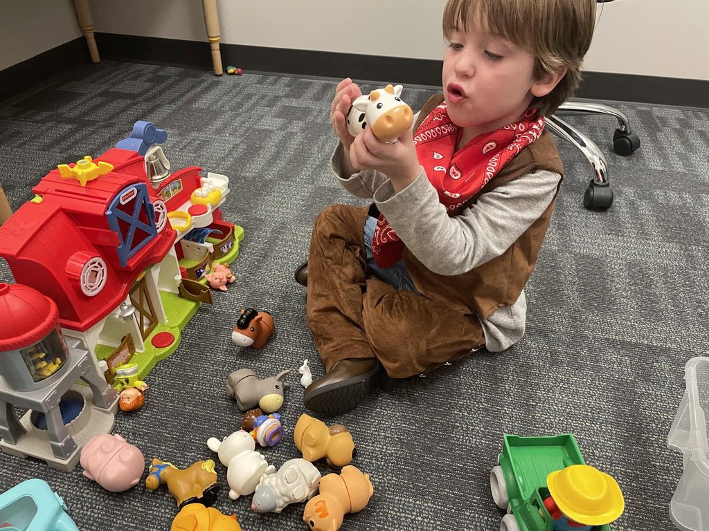 Toddler in a cowboy vest and red bandana holding a toy cow, mouth open mid-moo, next to a toy barn.